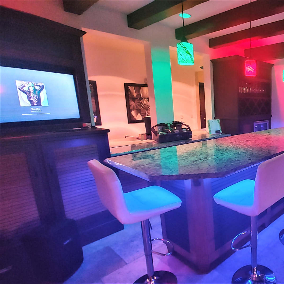 A modern kitchen bar area features a long granite countertop with high stools, accented by colorful pendant lights and a television in the background.