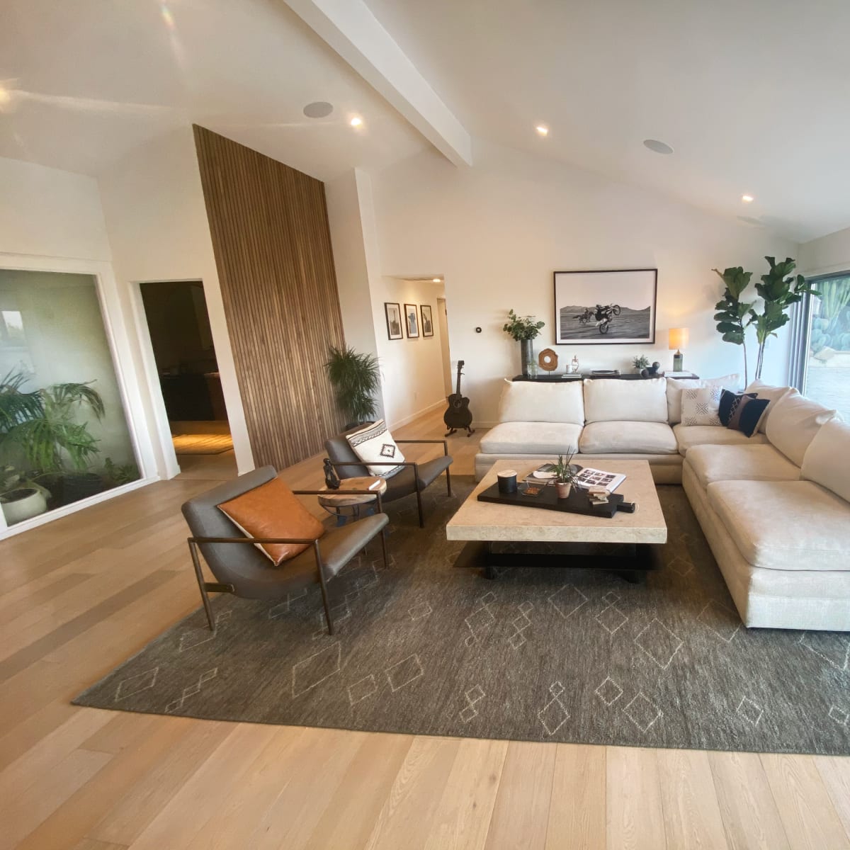 A modern living room with a large sectional sofa, a coffee table, and a pair of armchairs, surrounded by indoor plants and large windows that provide natural light.