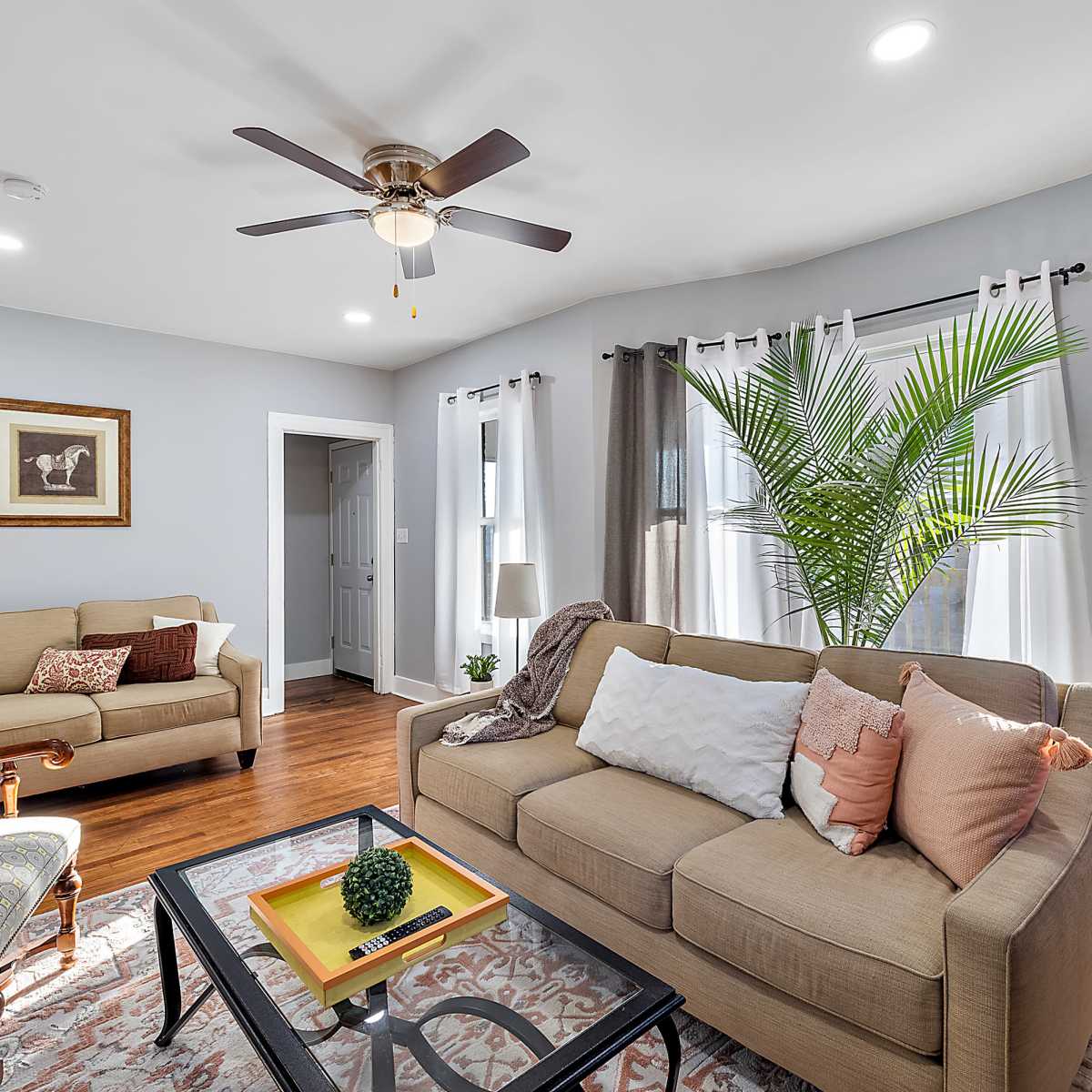 A well-lit living room features a sofa with decorative pillows, a coffee table with a plant, and a chair, surrounded by large windows and curtains.