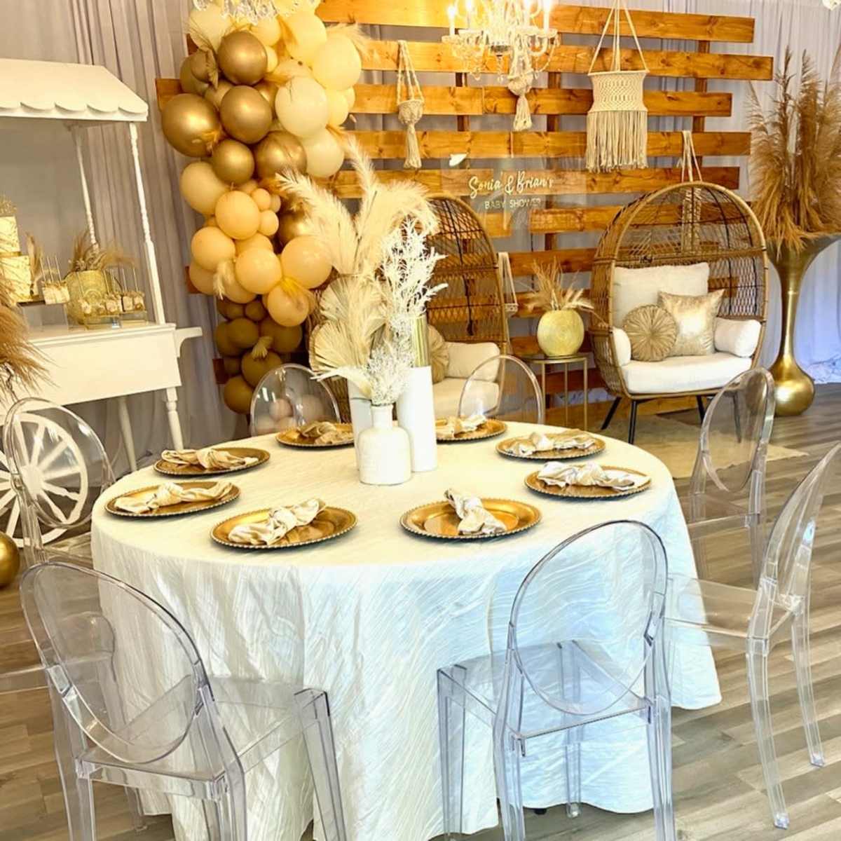 A decorated room with a round dining table set for a gathering, surrounded by clear acrylic chairs, and featuring balloons, a hanging chandelier, and a rustic wooden backdrop.