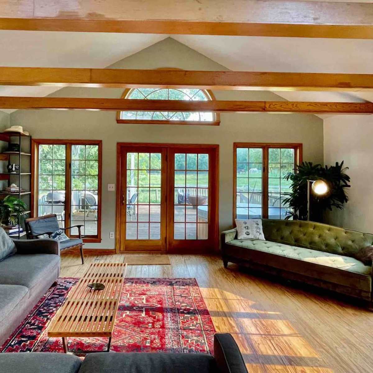 A bright living room with wooden beams, large windows, a gray sofa, a green tufted couch, a coffee table, and a patterned rug.