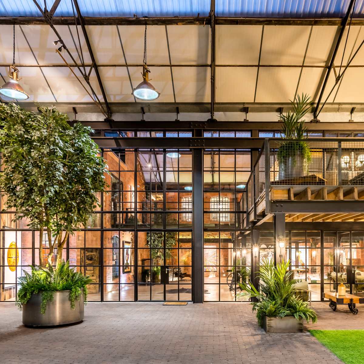 An expansive industrial-style interior with large glass windows, a mezzanine level, and greenery integrated into the space.