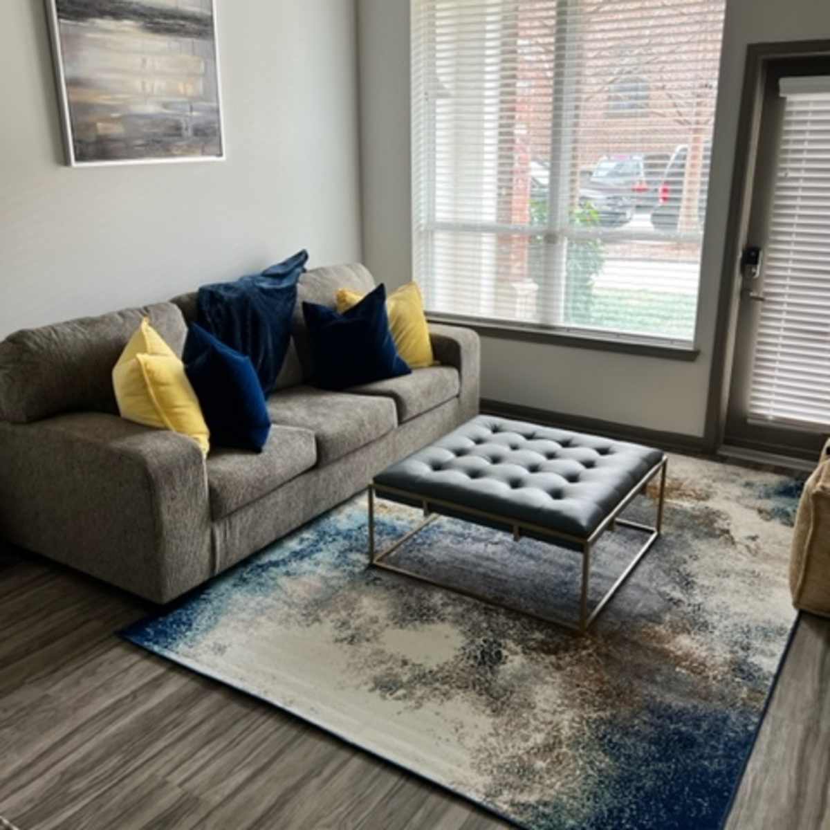 A living room with a grey sofa, two yellow and blue pillows, a tufted ottoman on a patterned rug, and large windows allowing natural light.