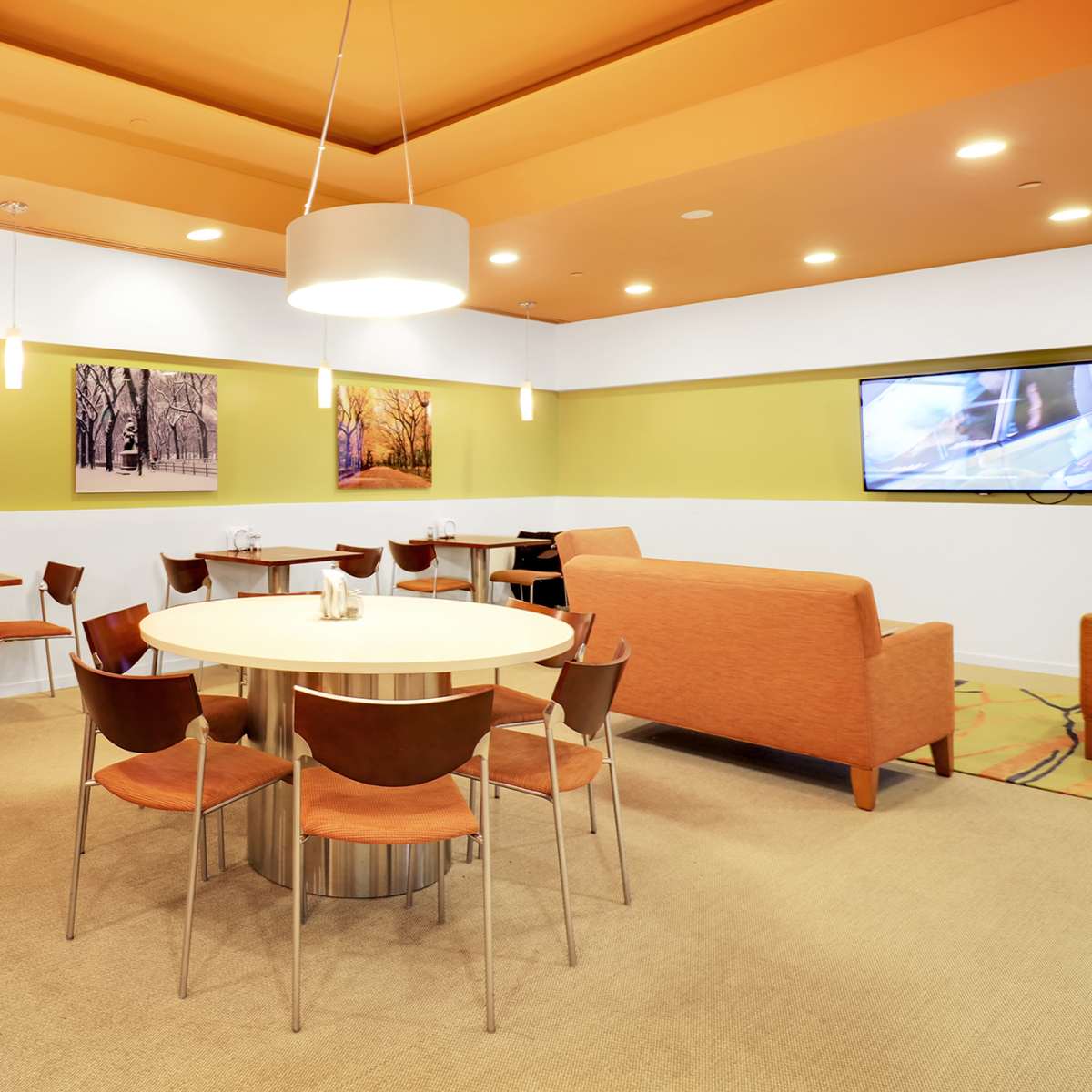 A modern break room featuring a round table surrounded by chairs, a sofa, and wall-mounted televisions, all in a brightly colored, well-lit space.