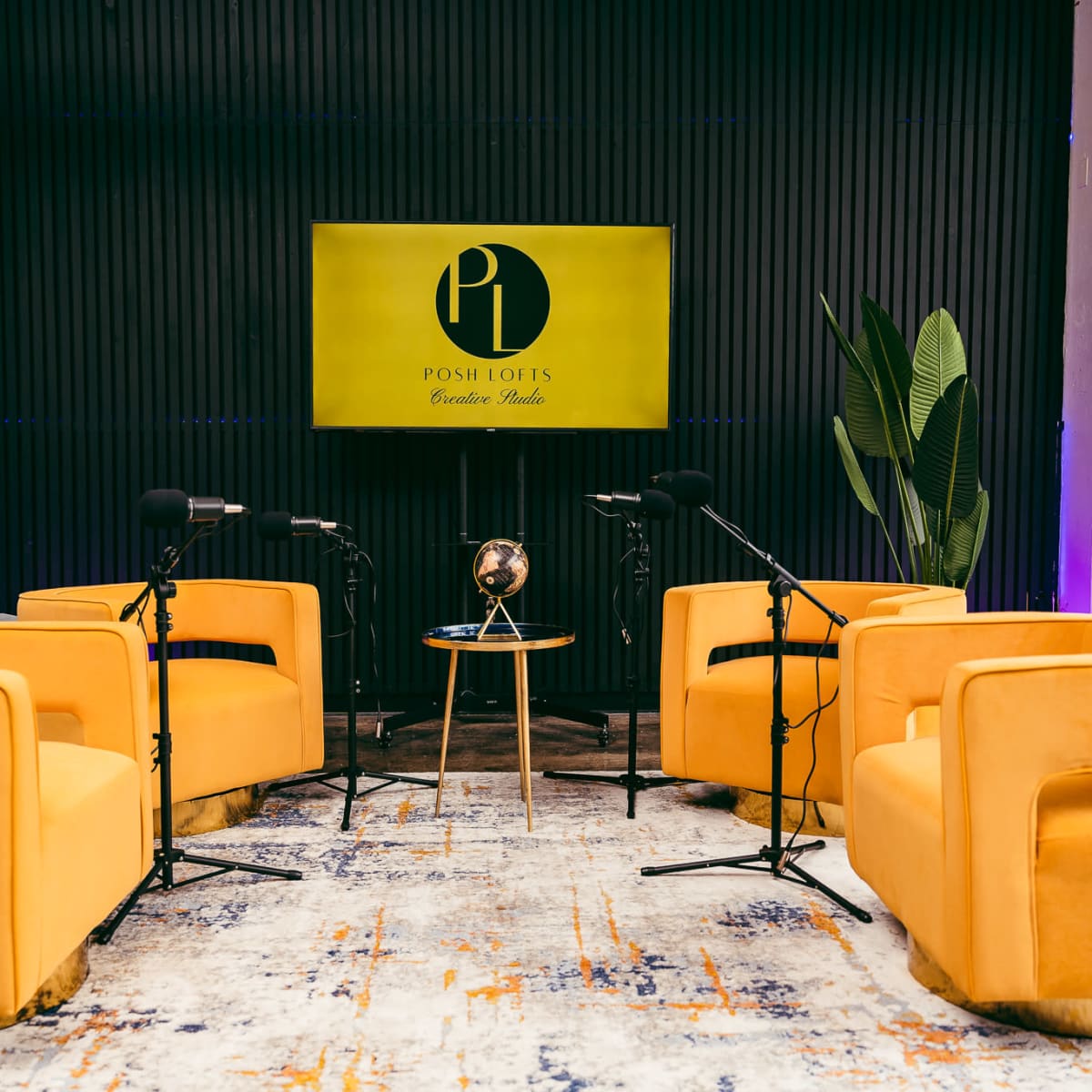 A modern interview setup features four yellow chairs arranged around a small table with a globe, positioned in front of a large screen displaying the logo "Posh Lotts Creative Studio."