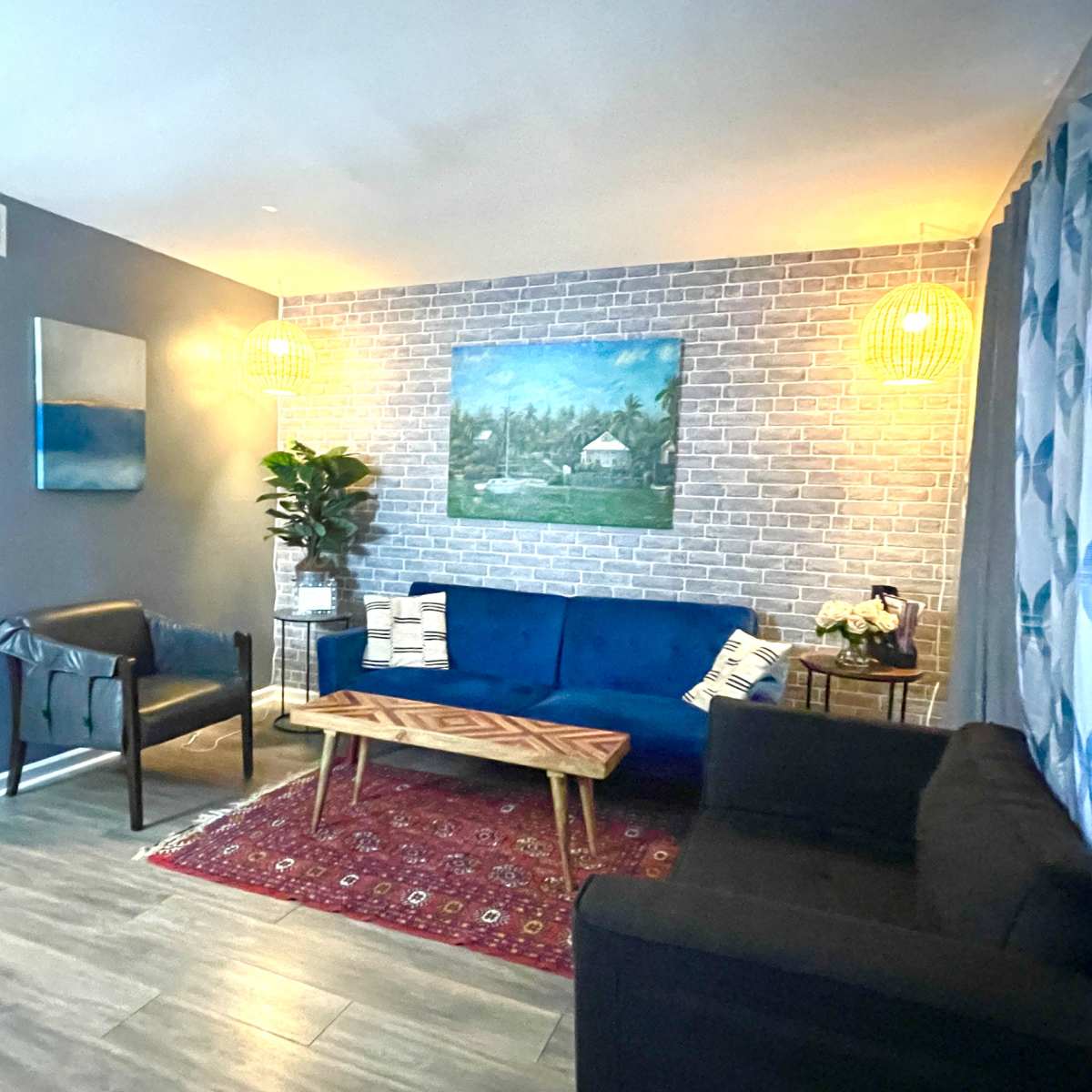 The living room features a blue velvet sofa and a dark couch, accented by a patterned area rug, a wooden coffee table, and a wall adorned with a landscape painting and decorative lighting.