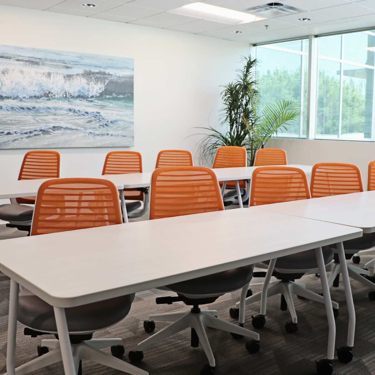 A bright, modern conference room featuring several white tables arranged in rows with orange mesh chairs and a large wave painting on the wall.