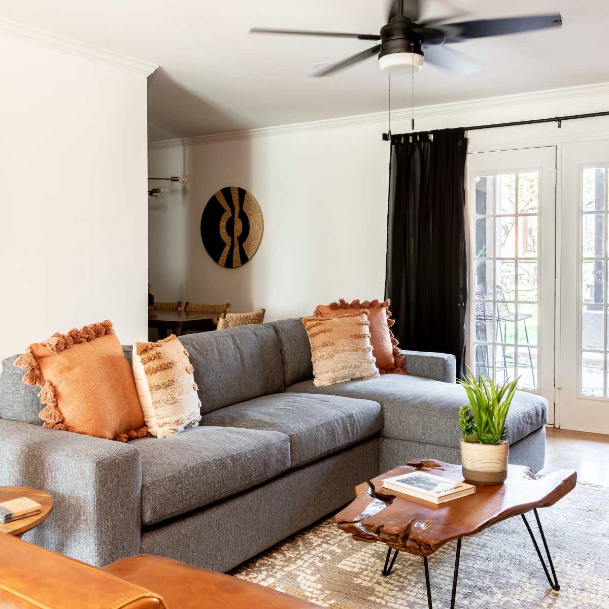 A modern living room features a gray sectional sofa with decorative pillows, a wooden coffee table, and large windows covered by black curtains.