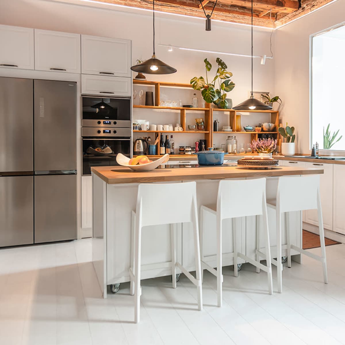 A modern kitchen featuring a gray refrigerator, white cabinetry, a wooden island with seating, and shelves displaying plants and kitchenware.