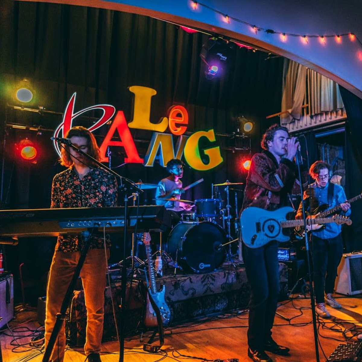 A band performs on stage with colorful lights and a backdrop displaying the word "BANG."