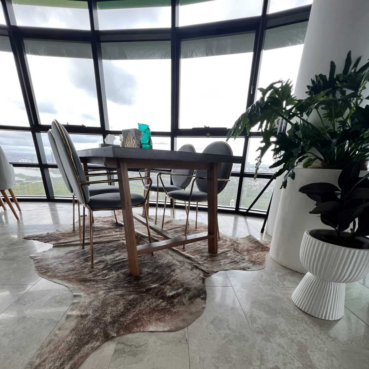 A modern dining area with a wooden table and chairs placed on a cowhide rug, surrounded by large windows that provide a view of the outside.
