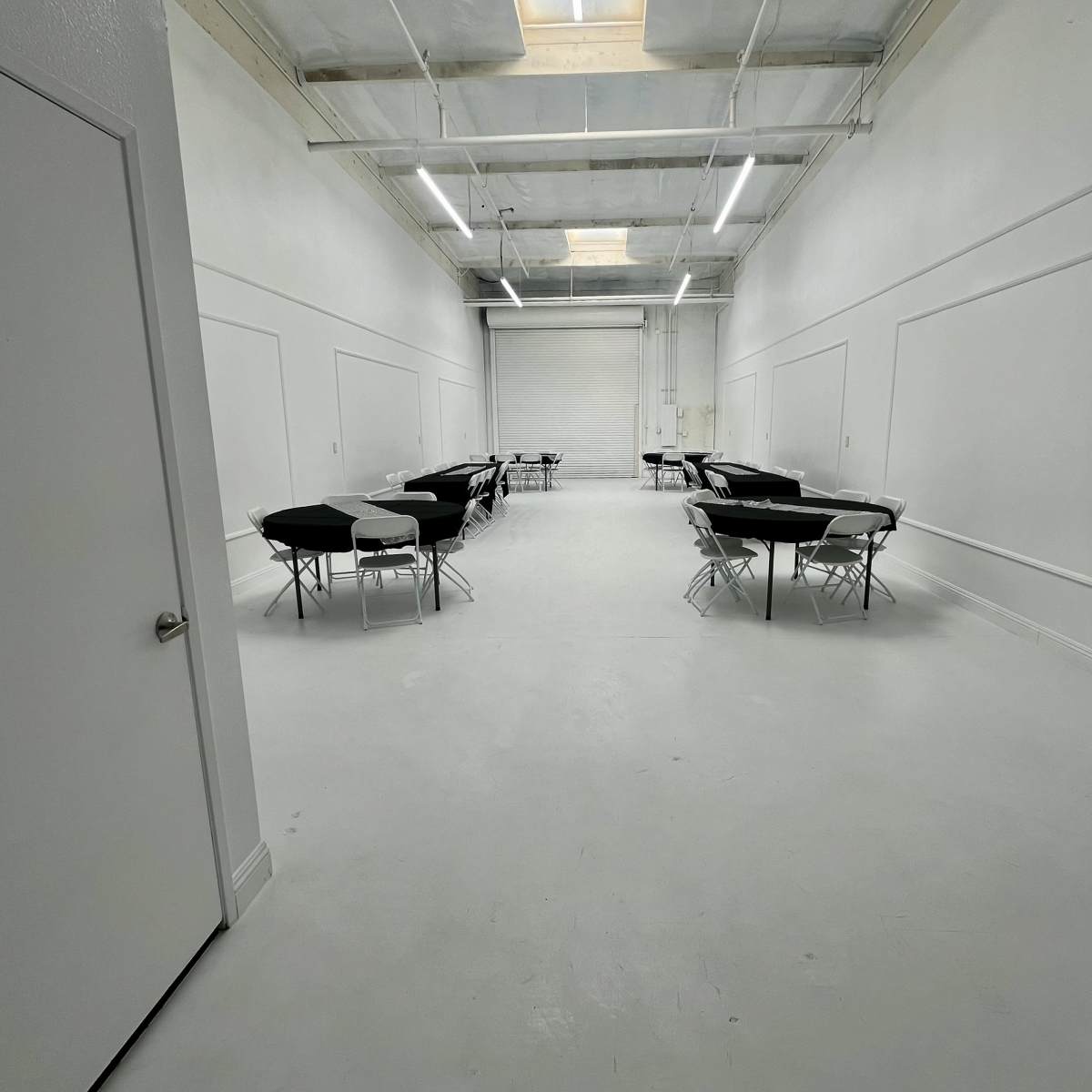 A large, empty room with white walls, a smooth white floor, and several black folding tables arranged in two rows.