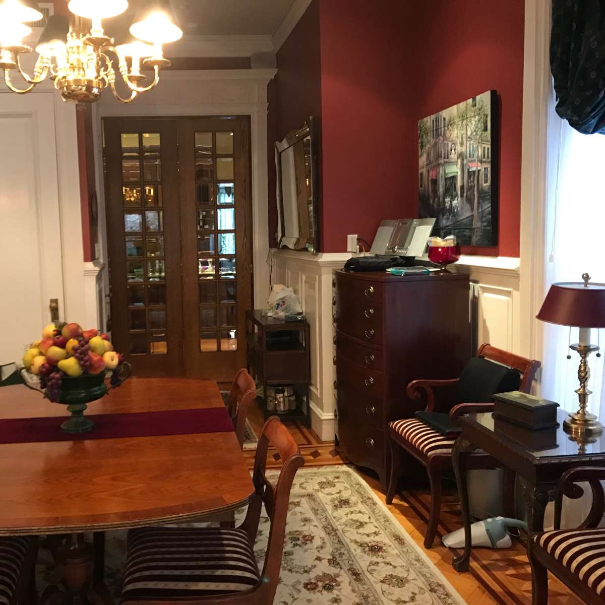 A dining room with a wooden table set for a meal, surrounded by chairs, and a large fruit bowl in the center, alongside a sideboard and a cabinet against the wall.