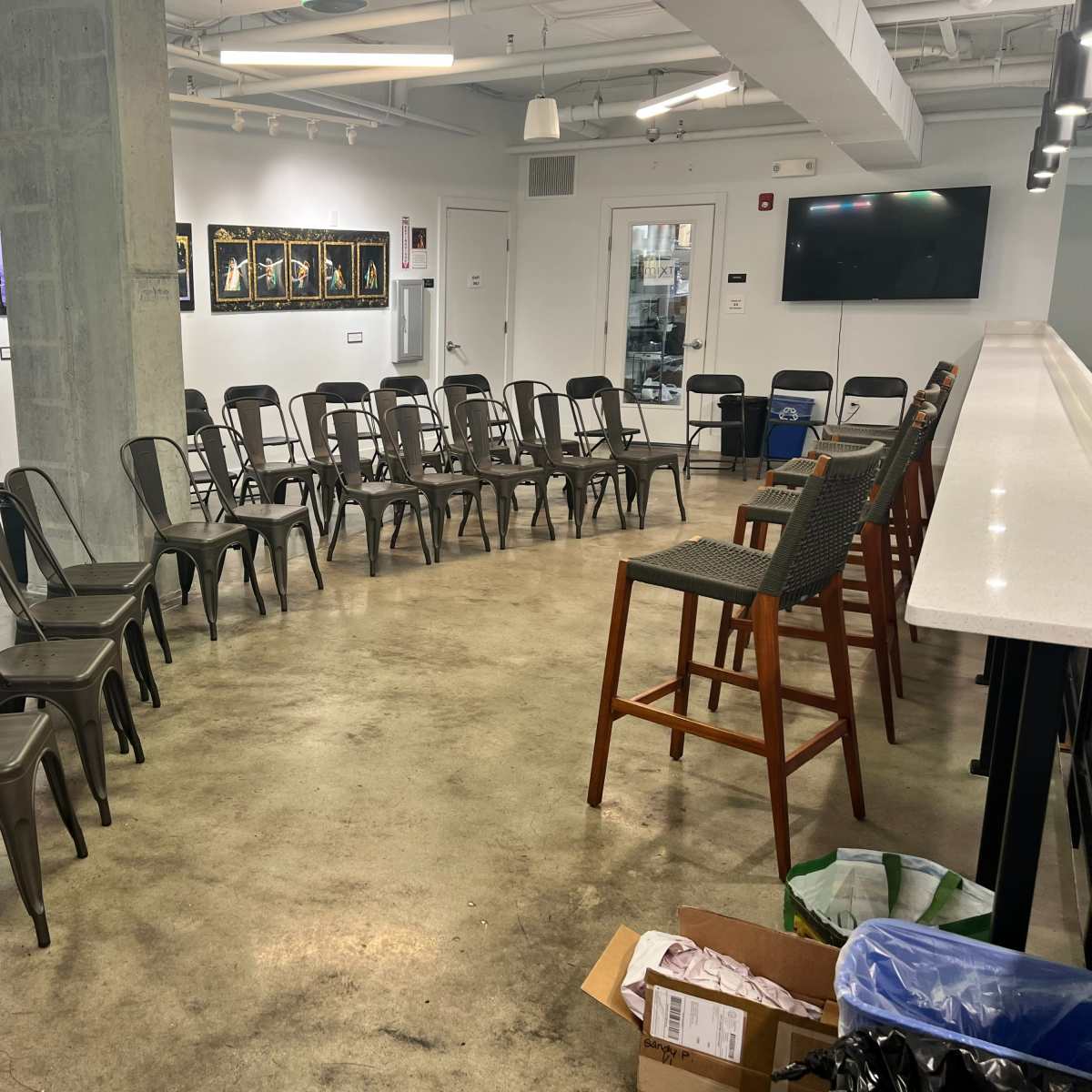 A room arranged with gray metal chairs in a circular formation around a central space, with a bar area and artwork displayed on the walls.