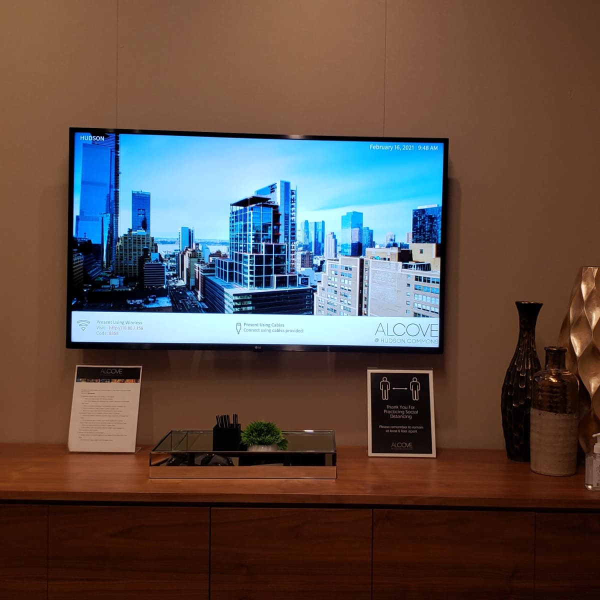 A modern room features a television displaying a cityscape, with a wooden console below that holds a plant, a tissue box, and decorative vases.
