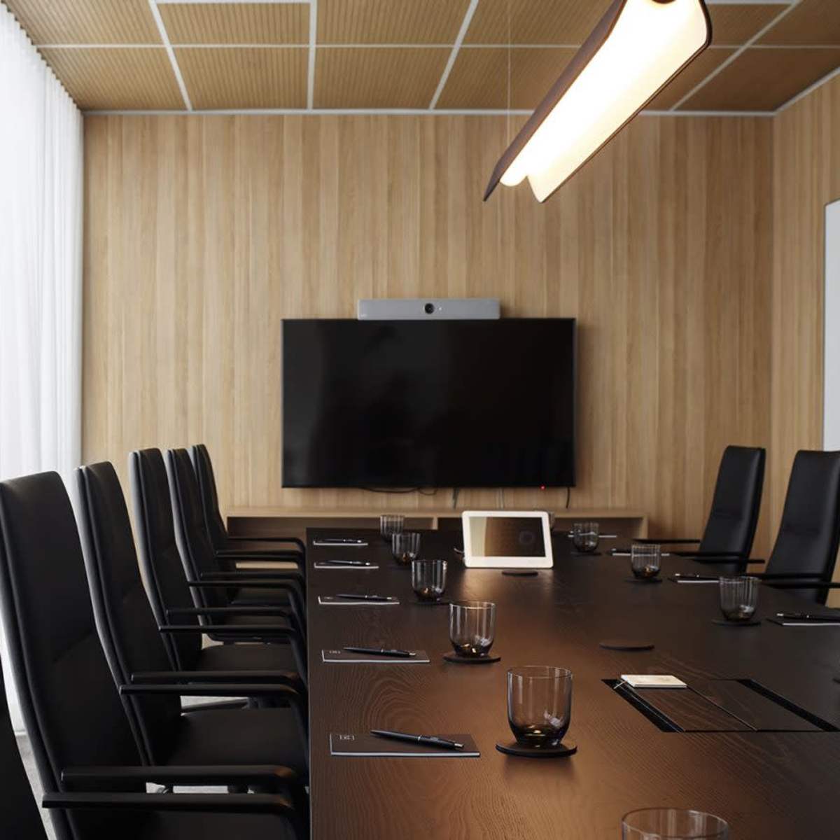 A conference room features a long table with black chairs, a large screen on the wall, and natural light filtering through sheer curtains.