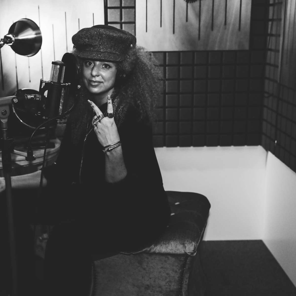 A person with curly hair wears a hat and gestures with their hand while seated in a recording studio.