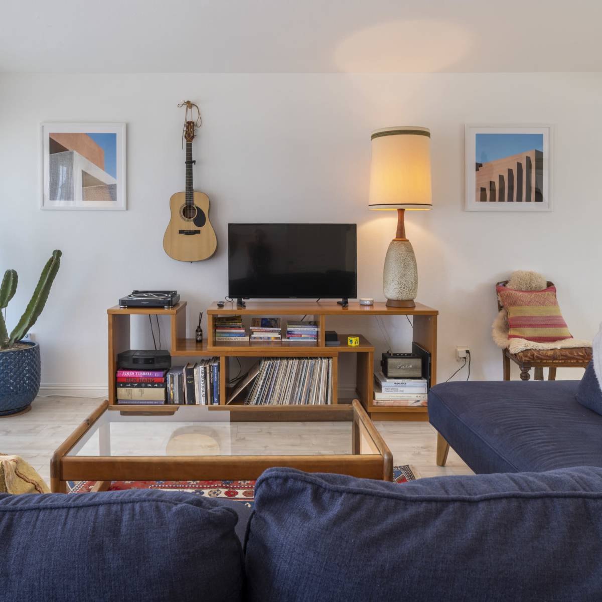 A living room features a navy blue sectional sofa, a coffee table with glass top, a wooden TV stand, and decor including a guitar, plant, and framed artwork.