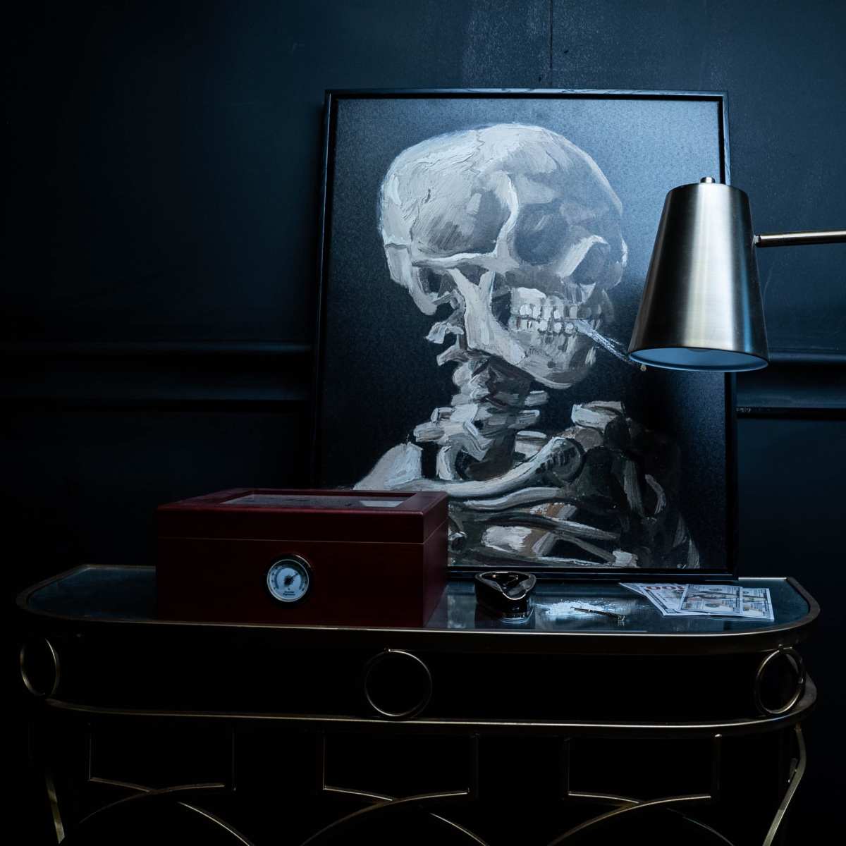A dark room features a framed painting of a skeleton on the wall, accompanied by a red box and a metallic lamp on a wooden table.