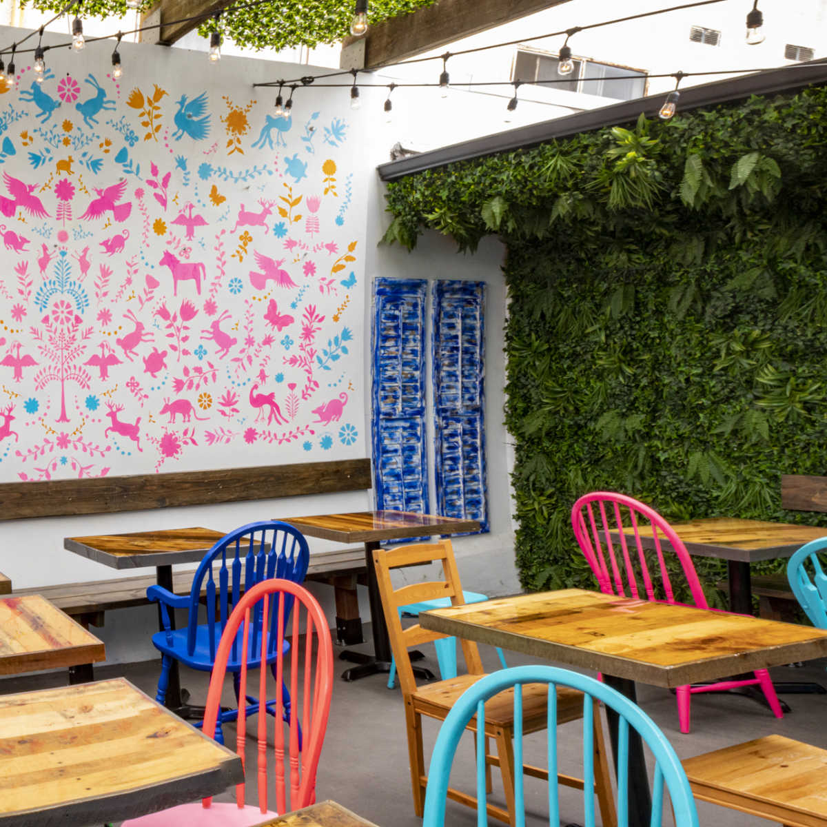 The outdoor seating area features wooden tables and brightly colored chairs against a backdrop of a vibrant mural and lush greenery.