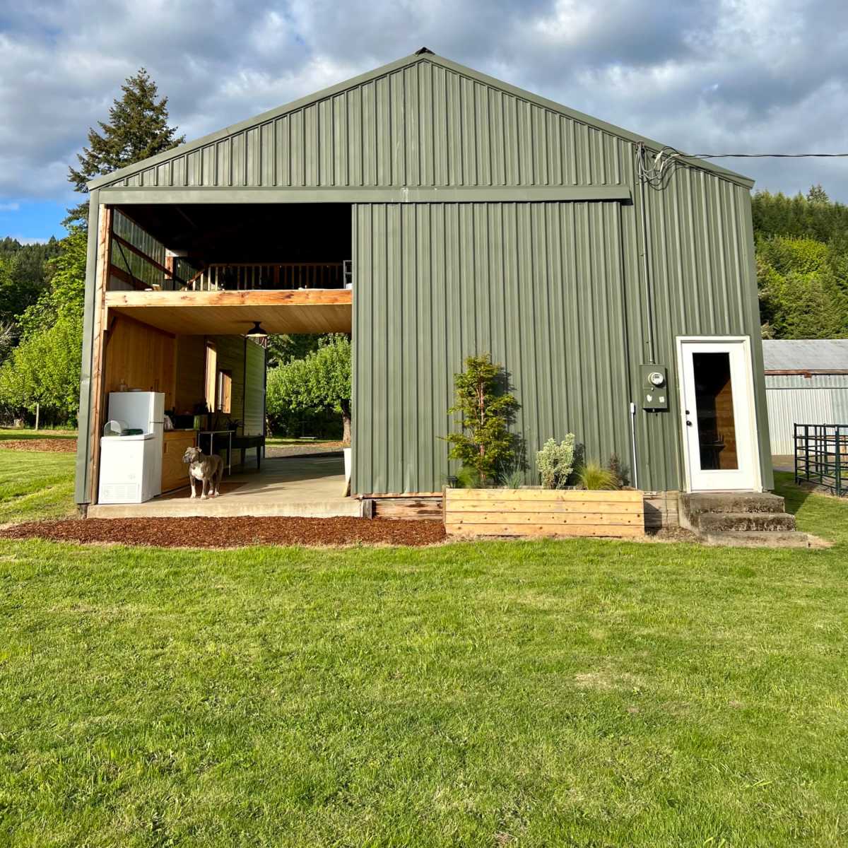 A large green metal barn features an open front with a wooden deck, set on a grassy landscape under a partly cloudy sky.