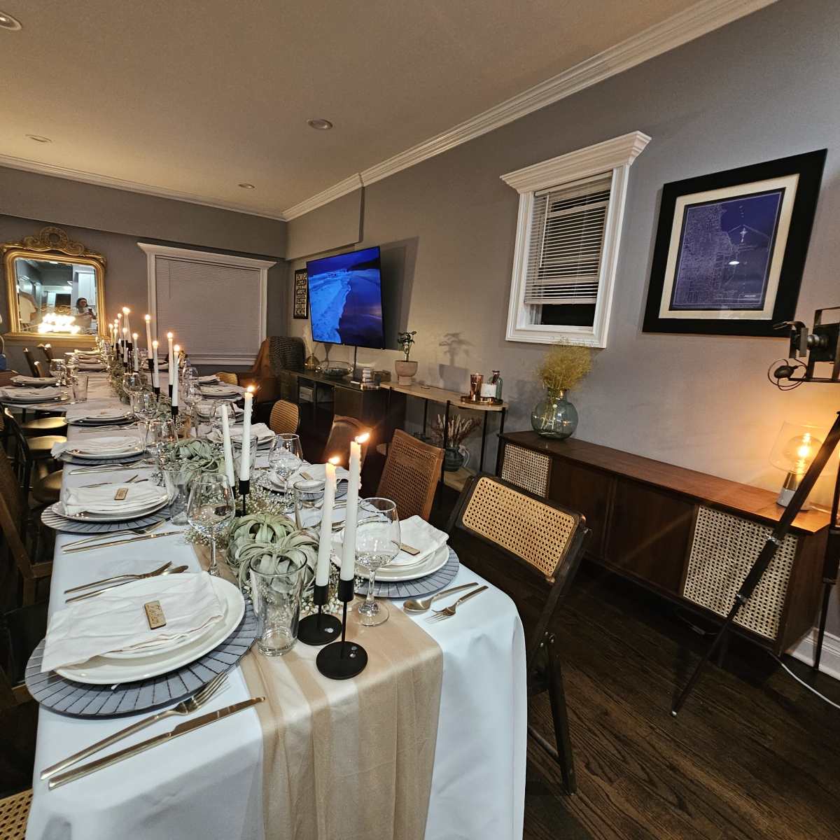 A long dining table is elegantly set with white plates, glowing candles, and decorative greenery, while a television displays a scenic image in a room with gray walls and wooden accents.