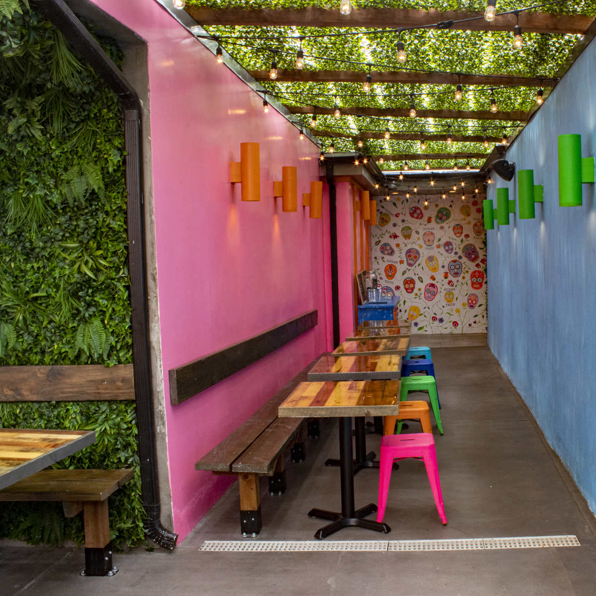 A brightly colored hallway features tables along one side and vibrant wall decorations, with green plants on the left and painted walls in pink and blue.