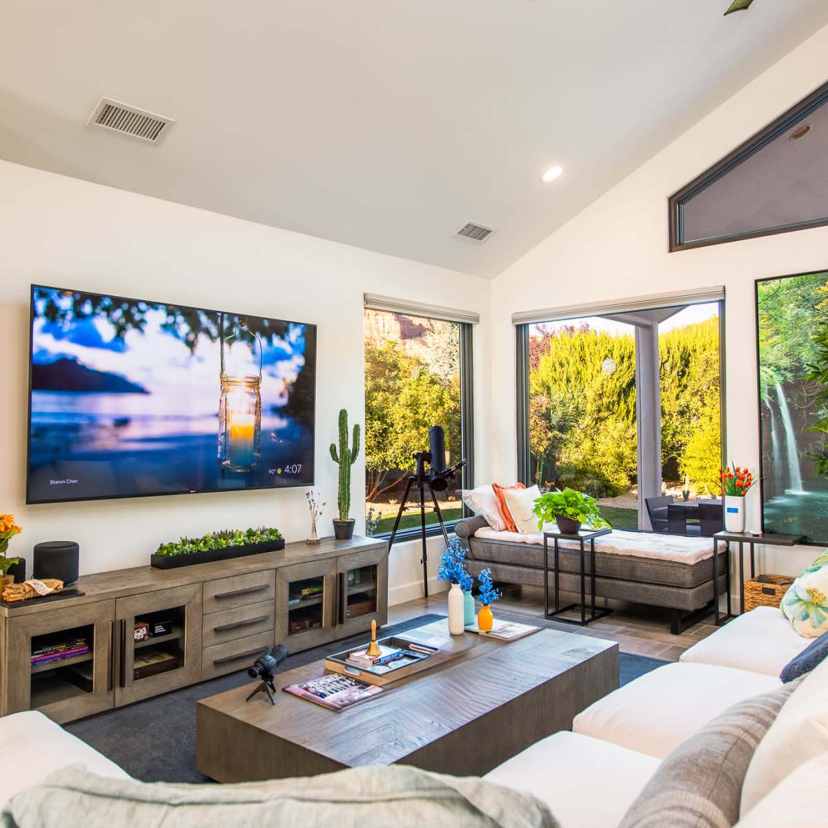 A modern living room with a large television, a coffee table, and windows overlooking a garden, all surrounded by potted plants.