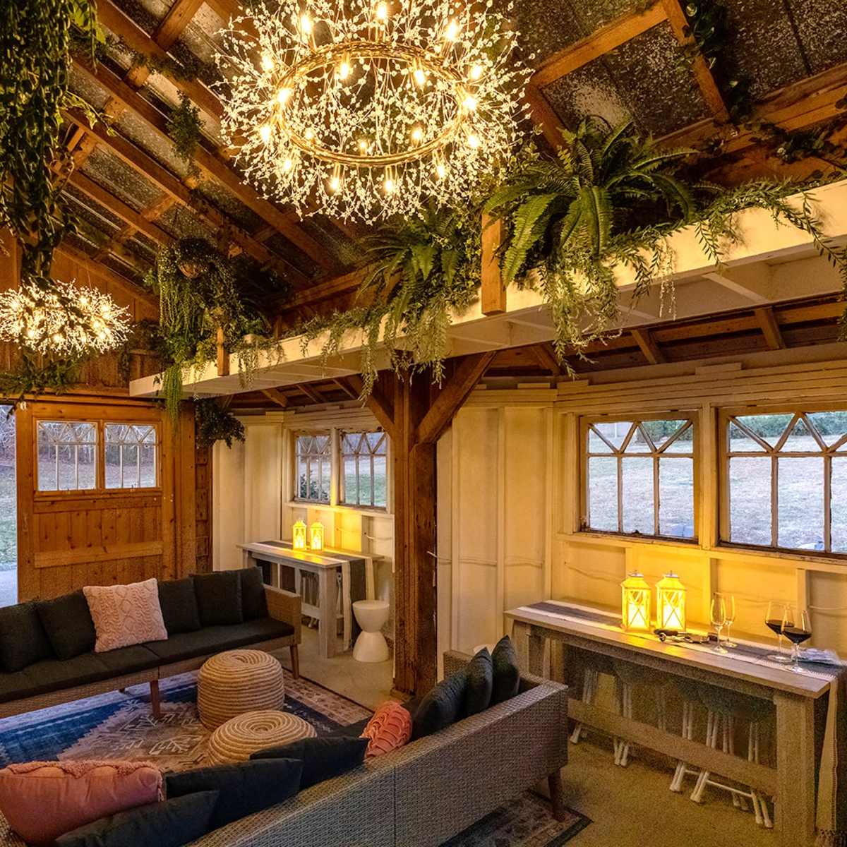 A rustic interior space with wooden beams, hanging greenery, and lit chandeliers, furnished with a cozy seating area and decorative lanterns.