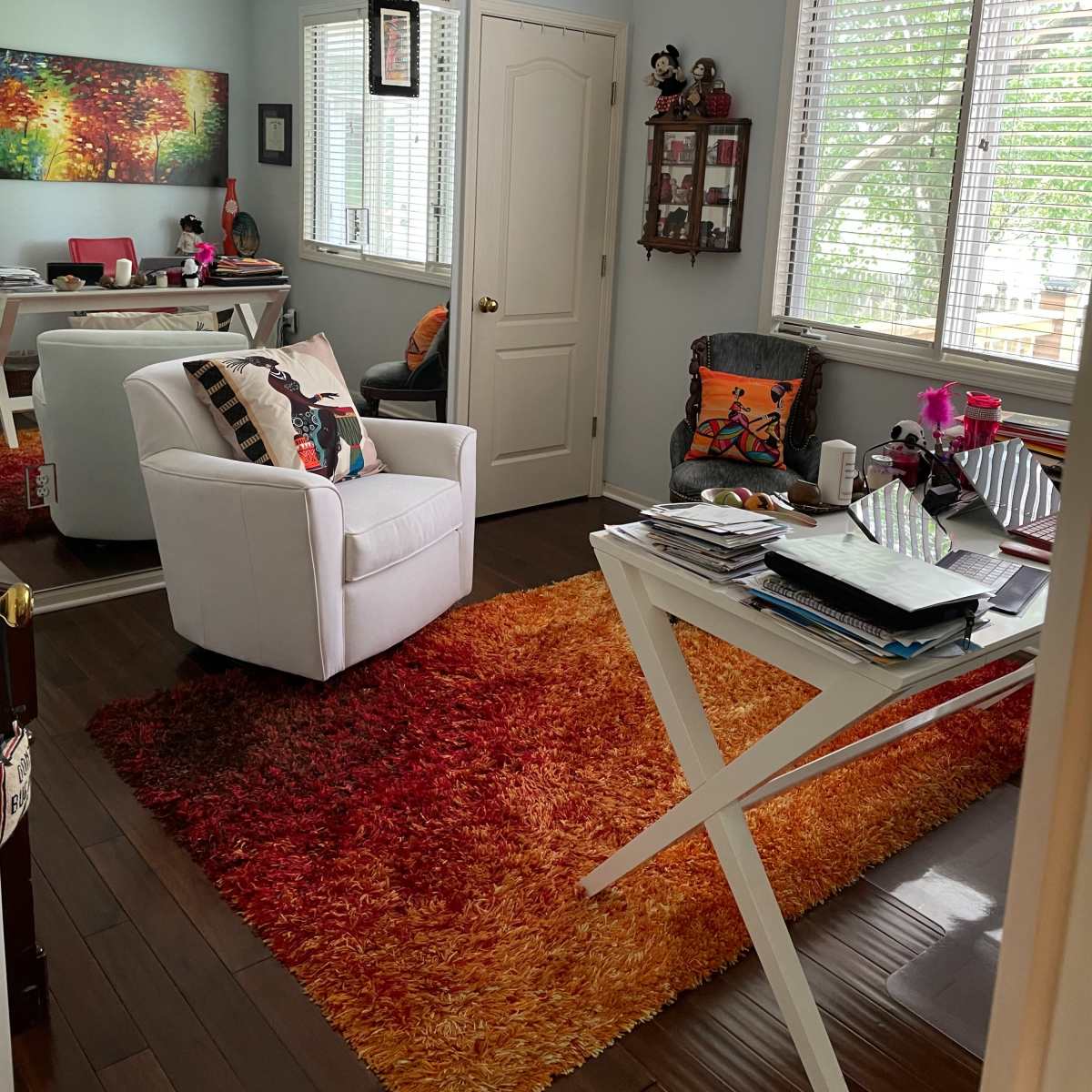 A brightly lit room with modern furnishings, including a white chair, a colorful area rug, a desk covered with papers, and windows letting in natural light.