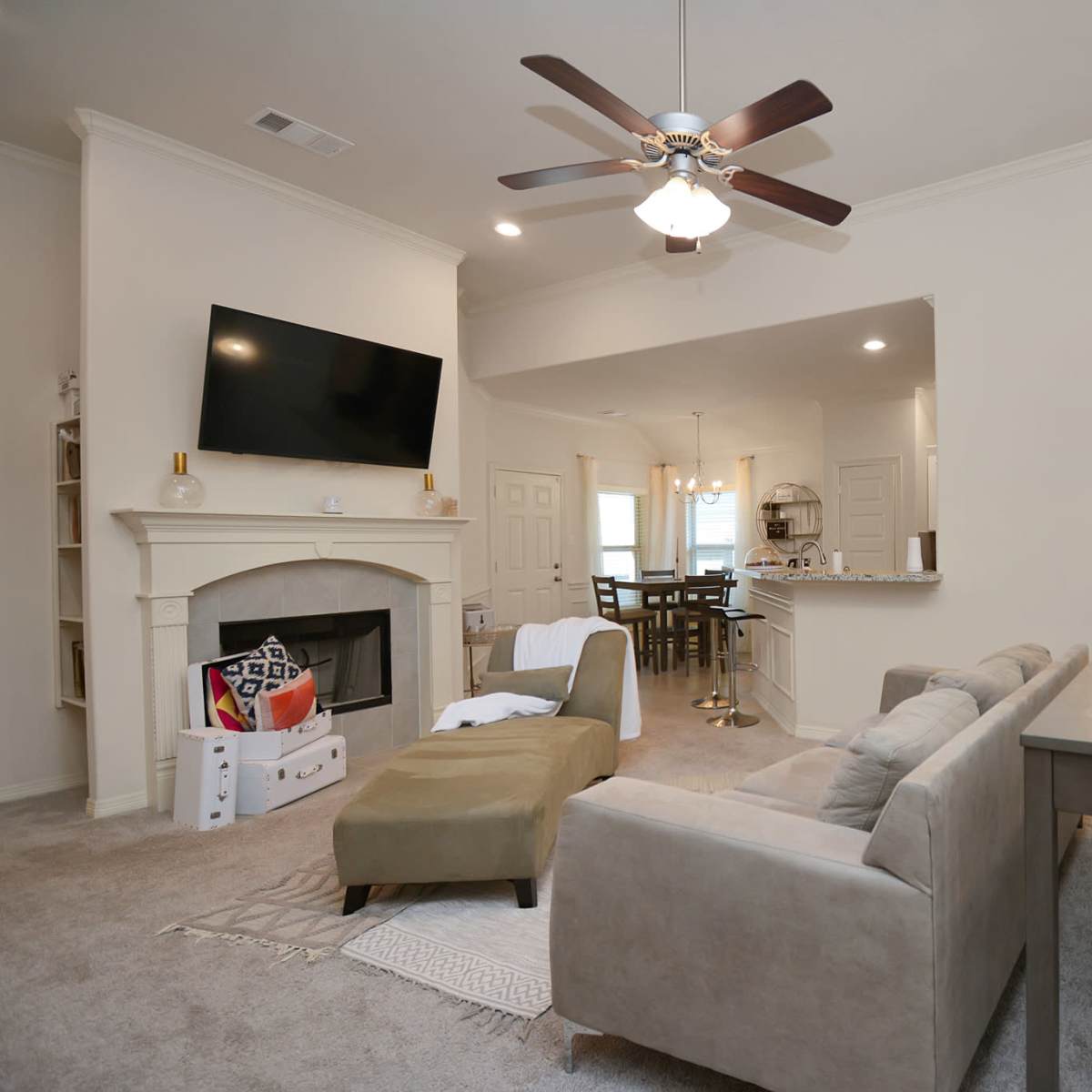 A living room with a beige sofa, a mounted television above a fireplace, and a dining area visible in the background.