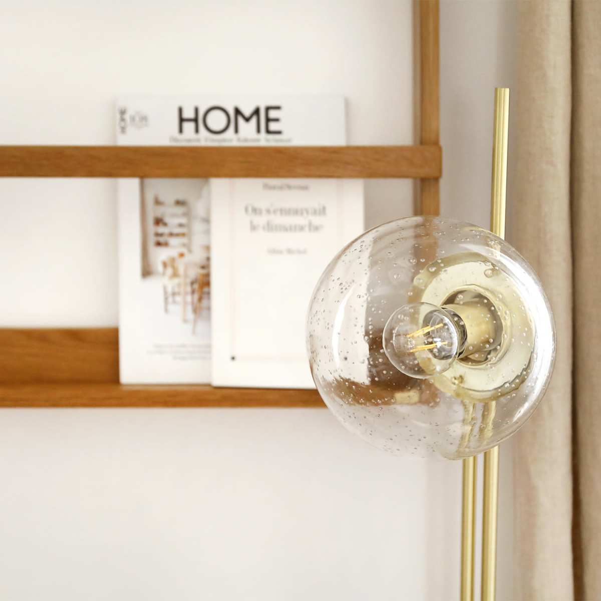 A clear glass lamp with a gold base stands in front of a wooden shelf displaying a magazine titled "HOME."