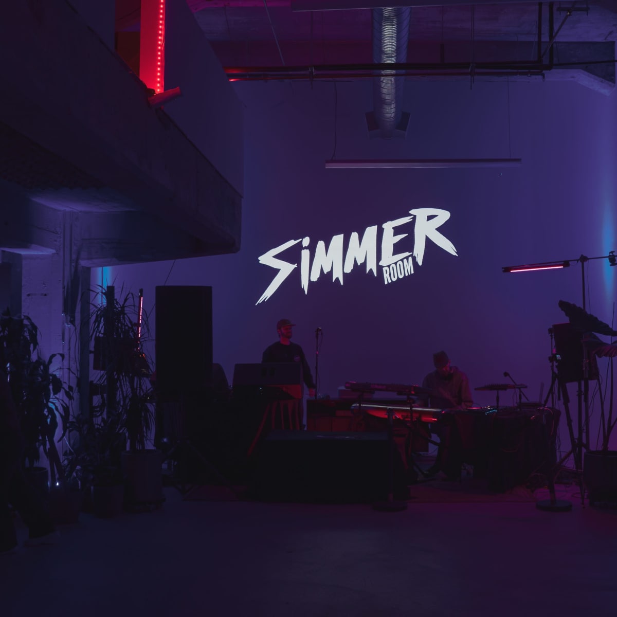 A dimly lit room with a large projection displaying the word "SIMMER," flanked by music equipment and decorative lighting.