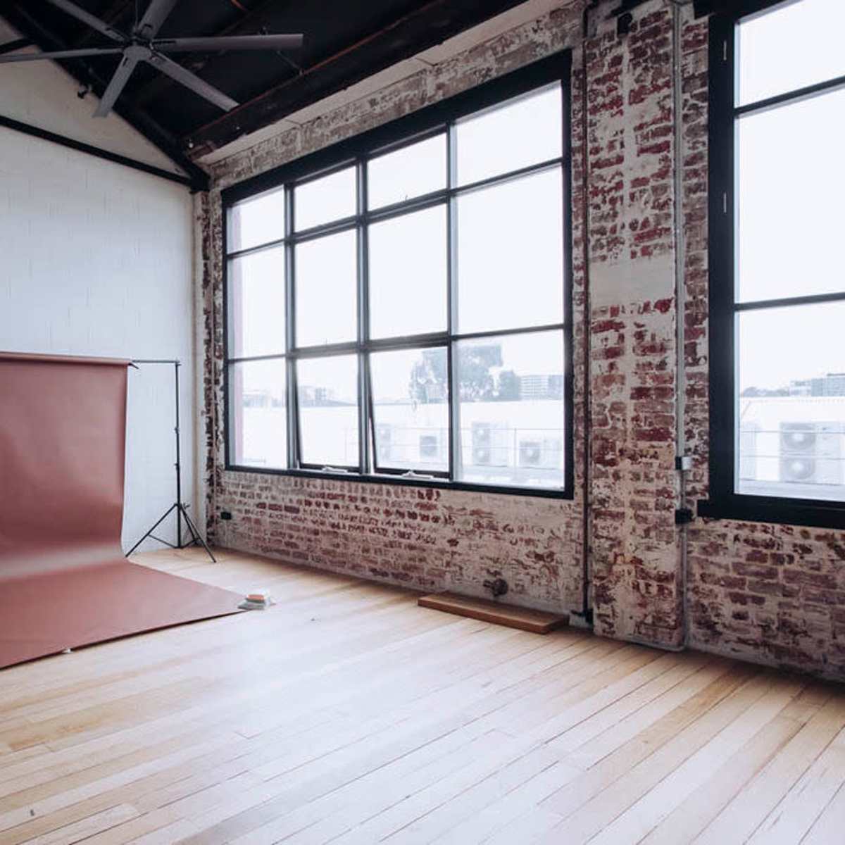 A spacious room with a brazen brick wall, large windows, and a brown backdrop set up on a wooden floor.