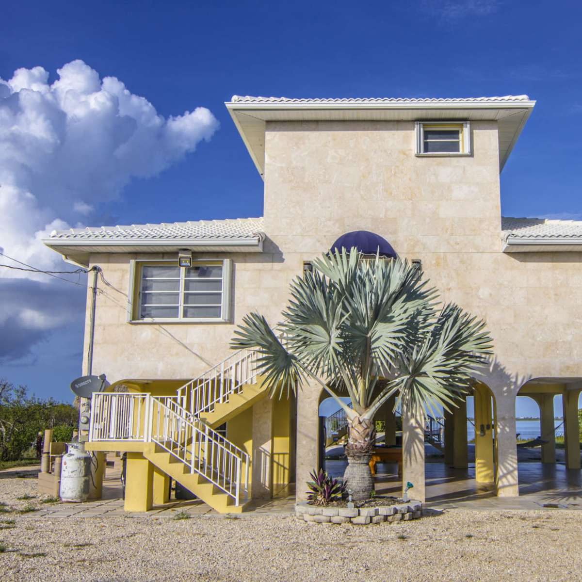 A two-story stone house with a palm tree in front and a staircase leading to the upper floor is set against a blue sky with clouds.