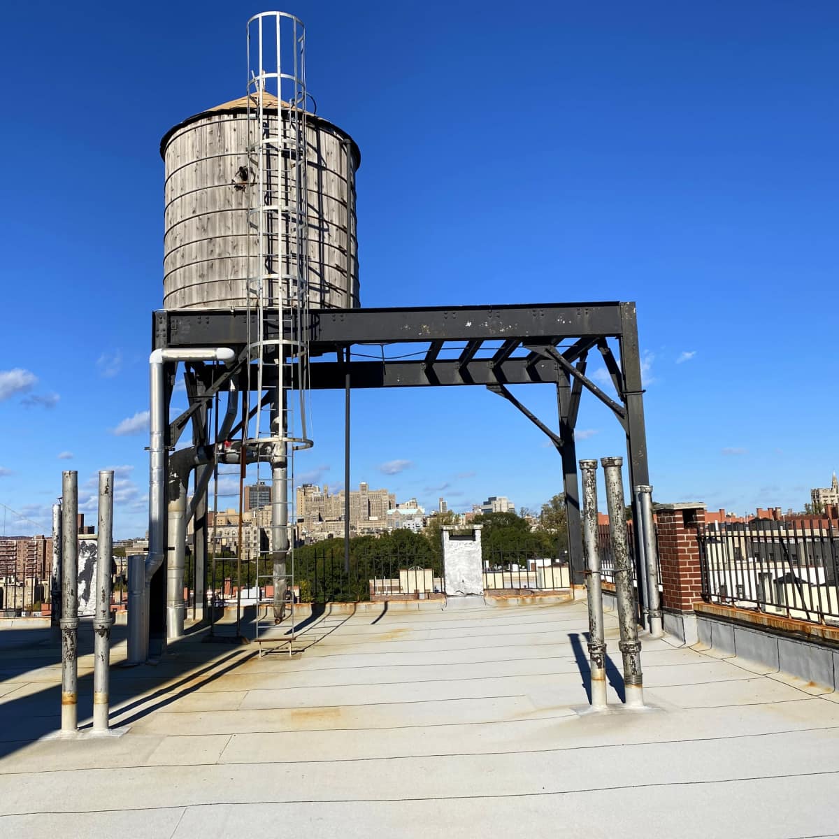 A wooden water tower stands on a rooftop with a clear blue sky and a city skyline in the background.