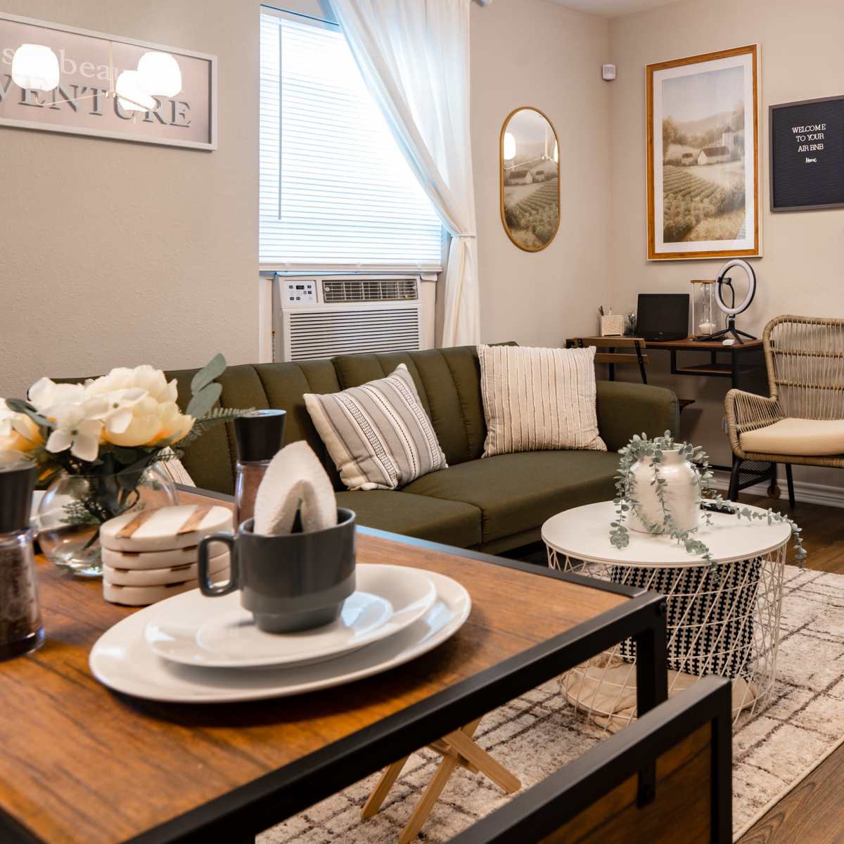 A cozy living room features a green sofa, two wicker chairs, a coffee table with decorative items, and large windows allowing natural light.