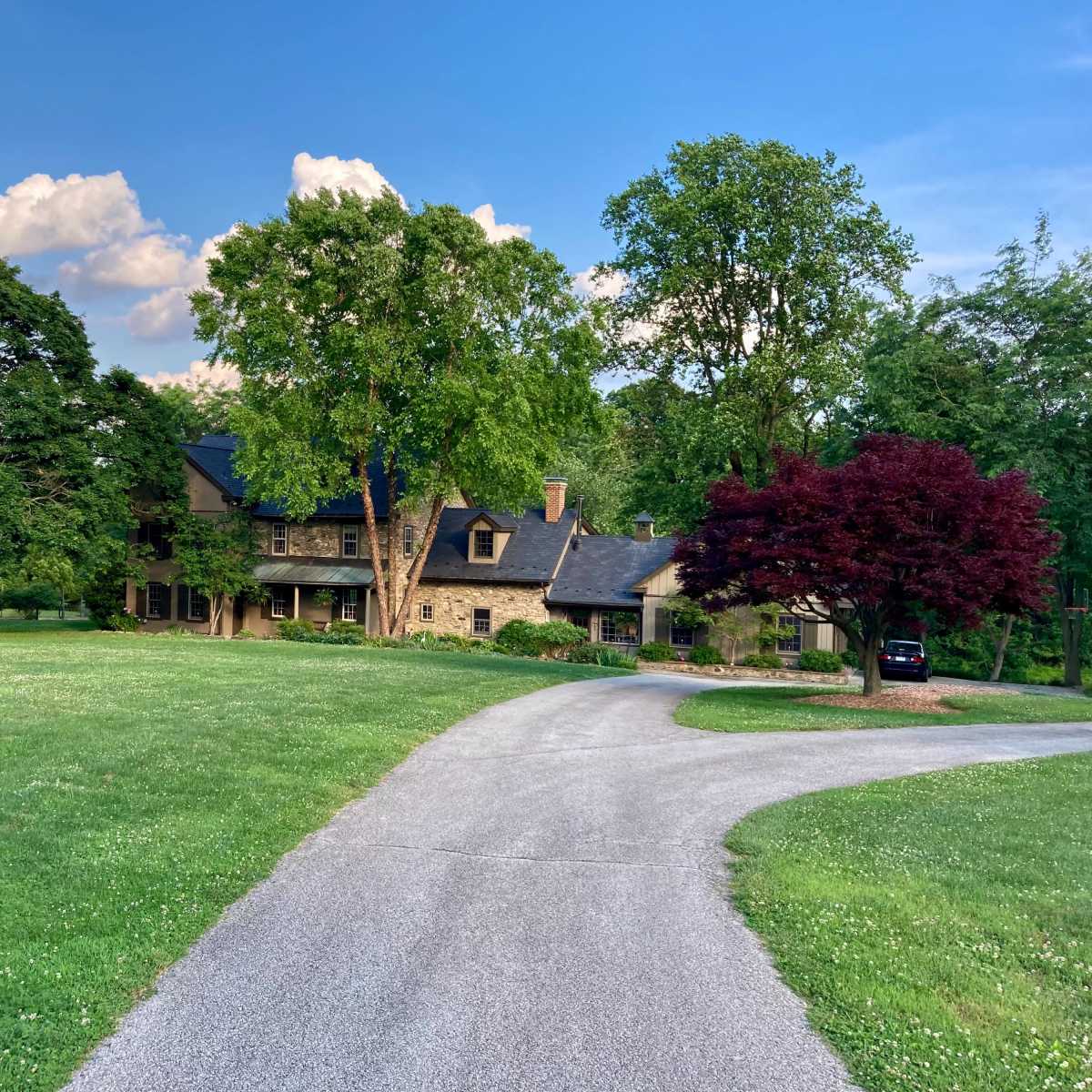 A large stone house with multiple trees and a curved driveway is set in a grassy landscape.