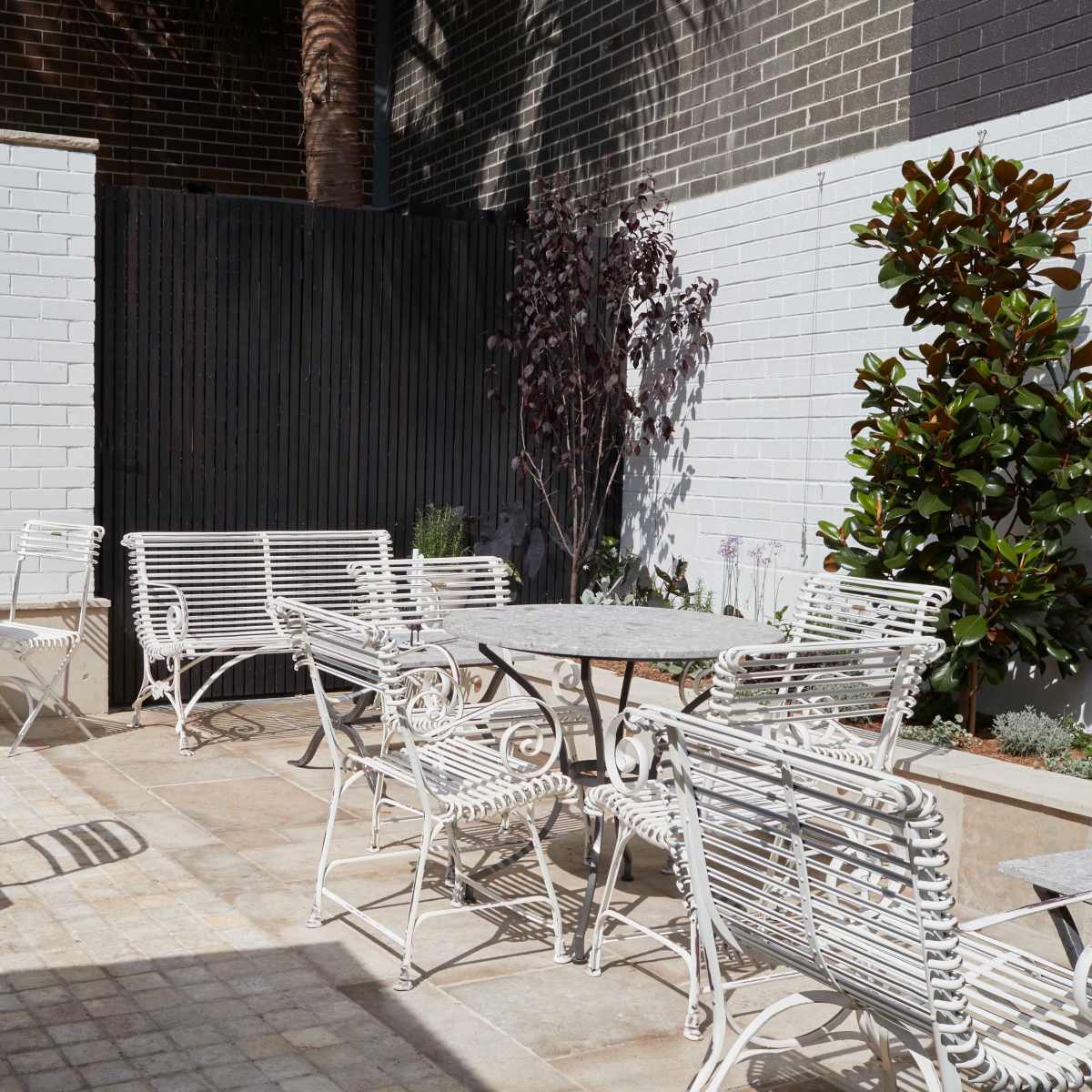 A courtyard features multiple white metal tables and chairs surrounded by greenery and textured walls.