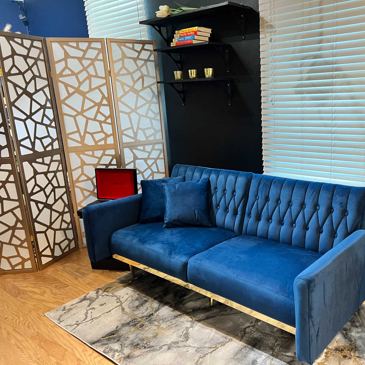 A blue velvet sofa with tufted upholstery, positioned near a decorative screen, shelves with books, and a side table with flowers.