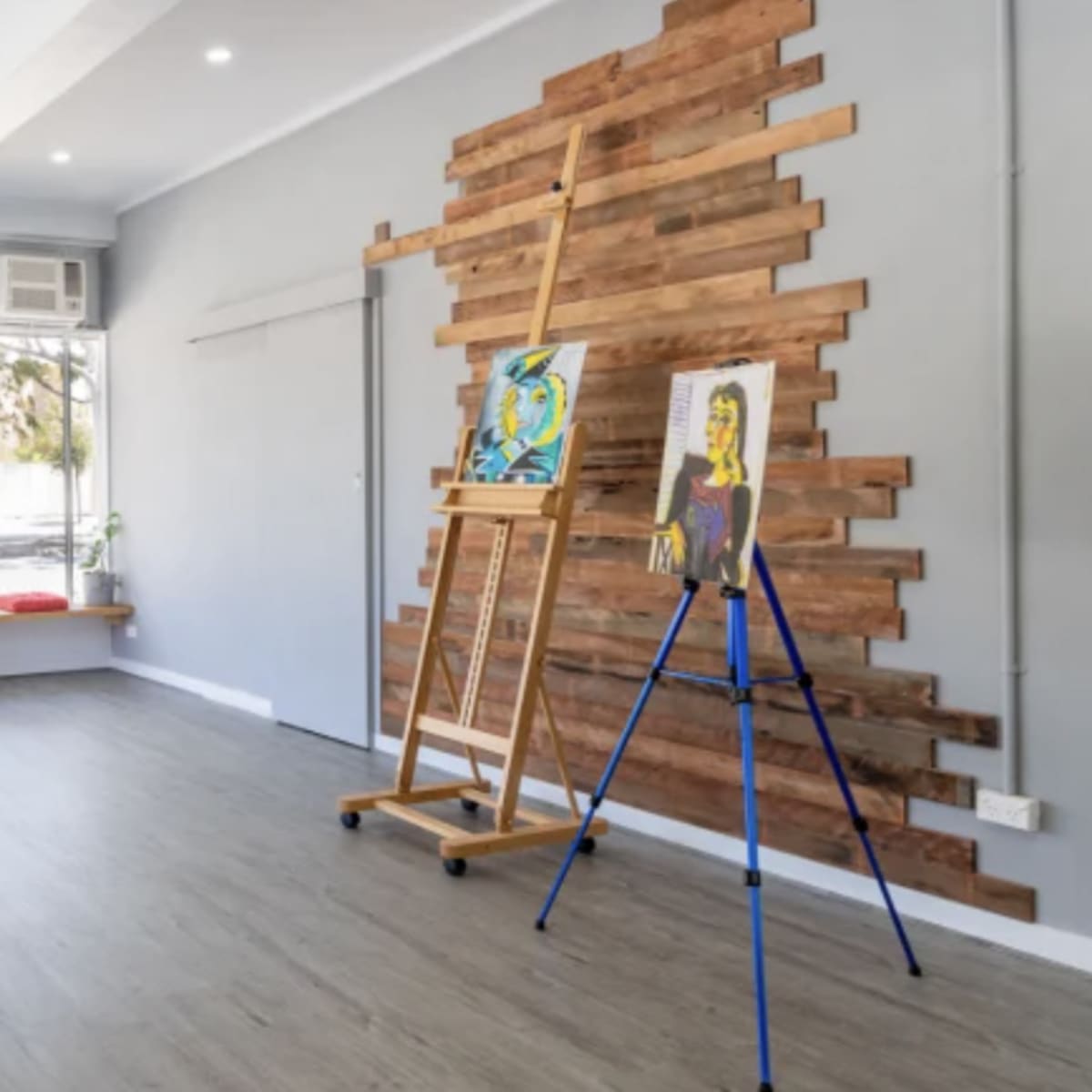 The interior of a spacious, well-lit art studio features wooden accents on the wall, an easel, and colorful cushions along the windowsill.