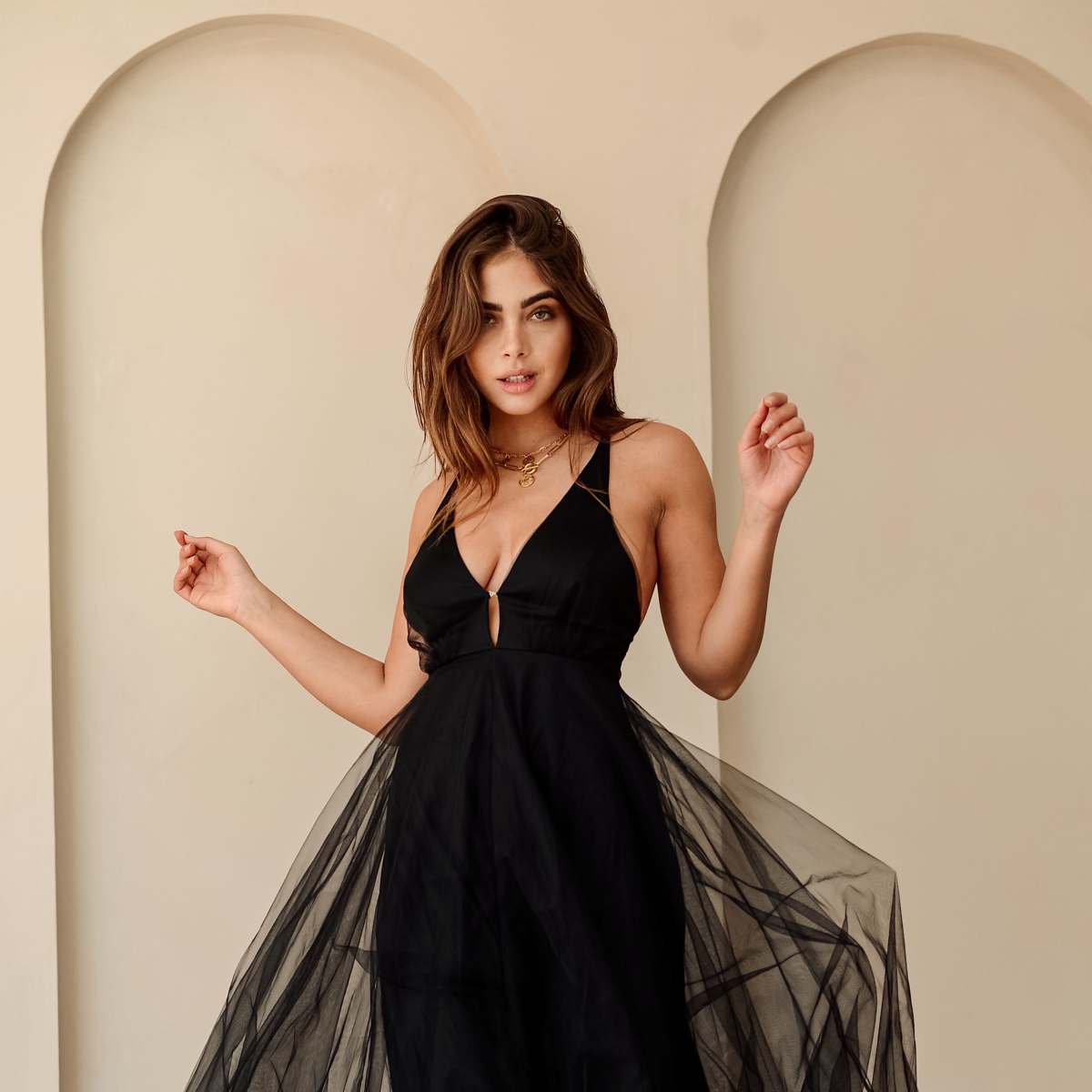 A woman poses in a black dress with a sheer overlay against a neutral background.
