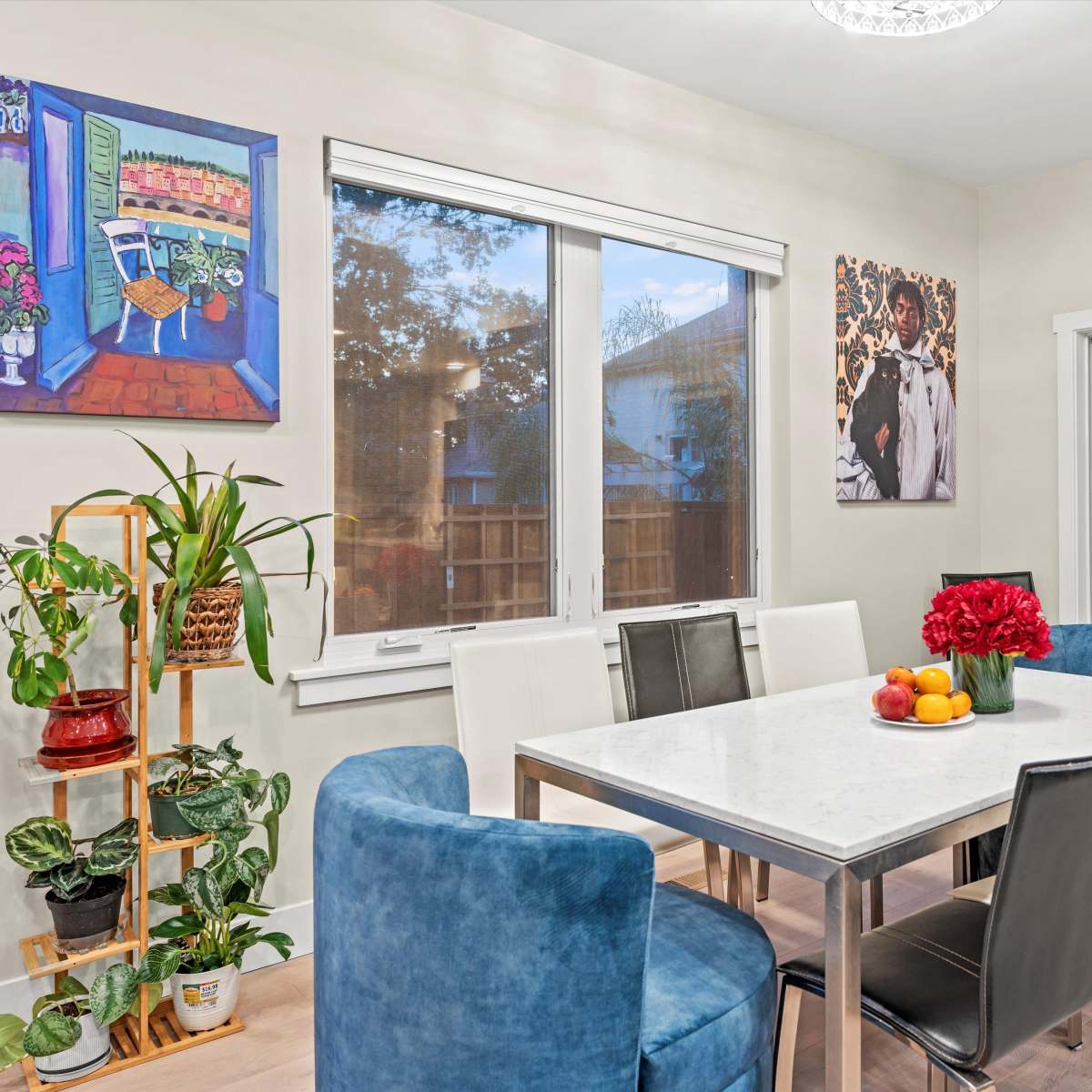 A dining area features a white table surrounded by colorful chairs, with large windows letting in natural light and plants adding greenery.