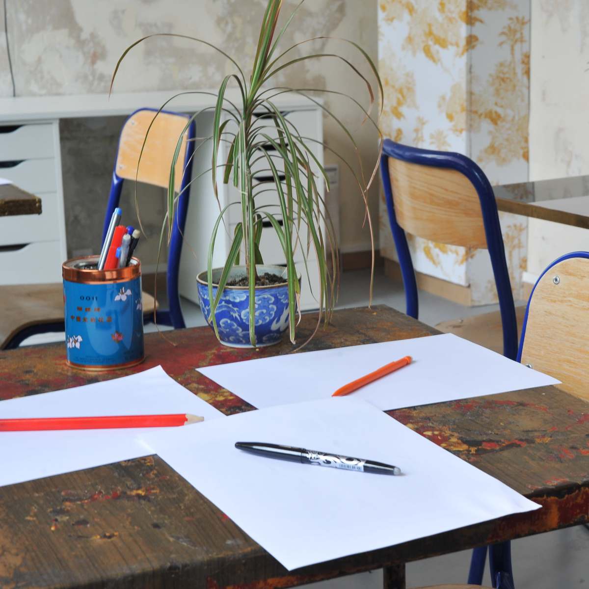 A wooden table with blank sheets of paper and colored pens is set in a room with chairs and potted plants.