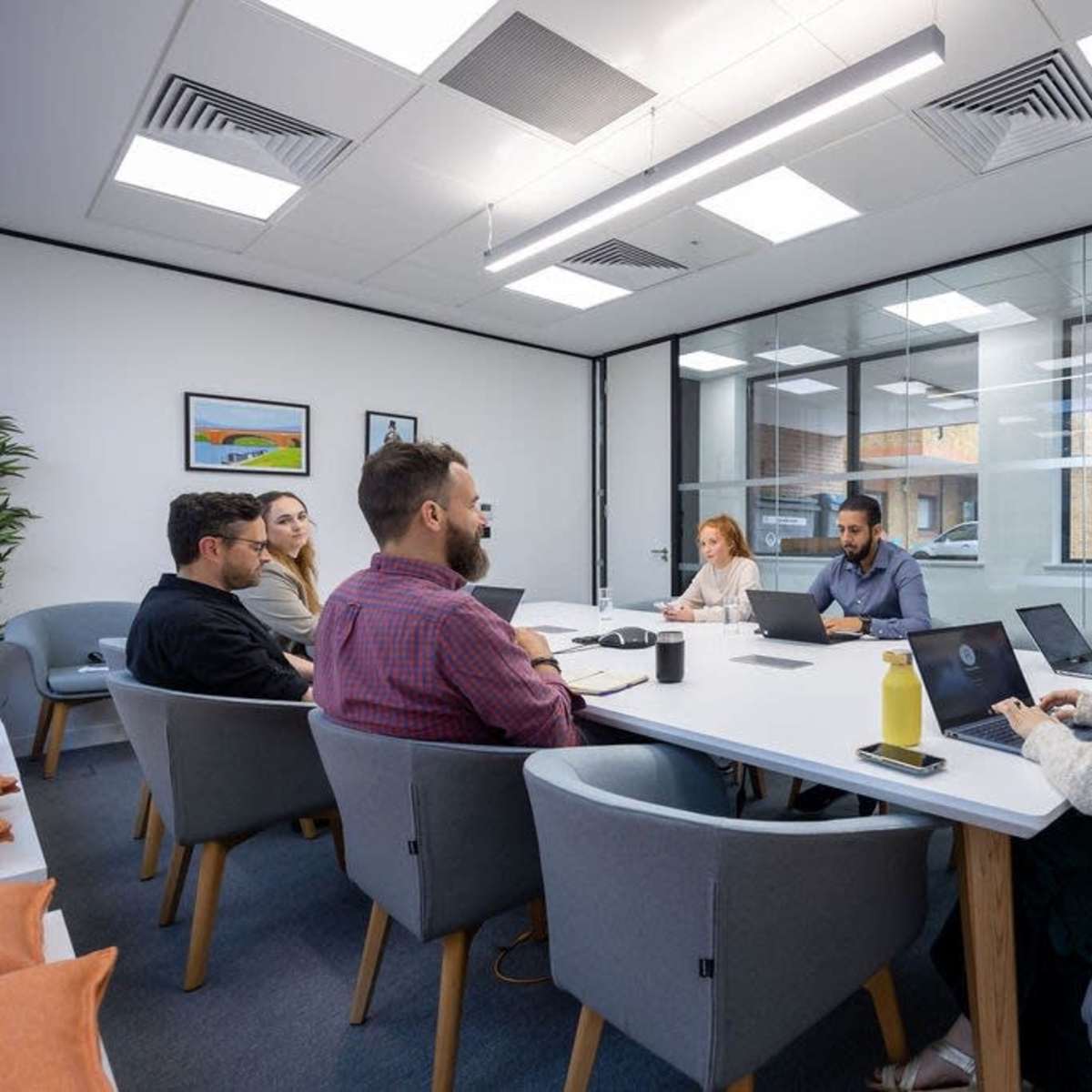 A group of six people sit around a large conference table in a well-lit meeting room, working on laptops and discussing ideas.