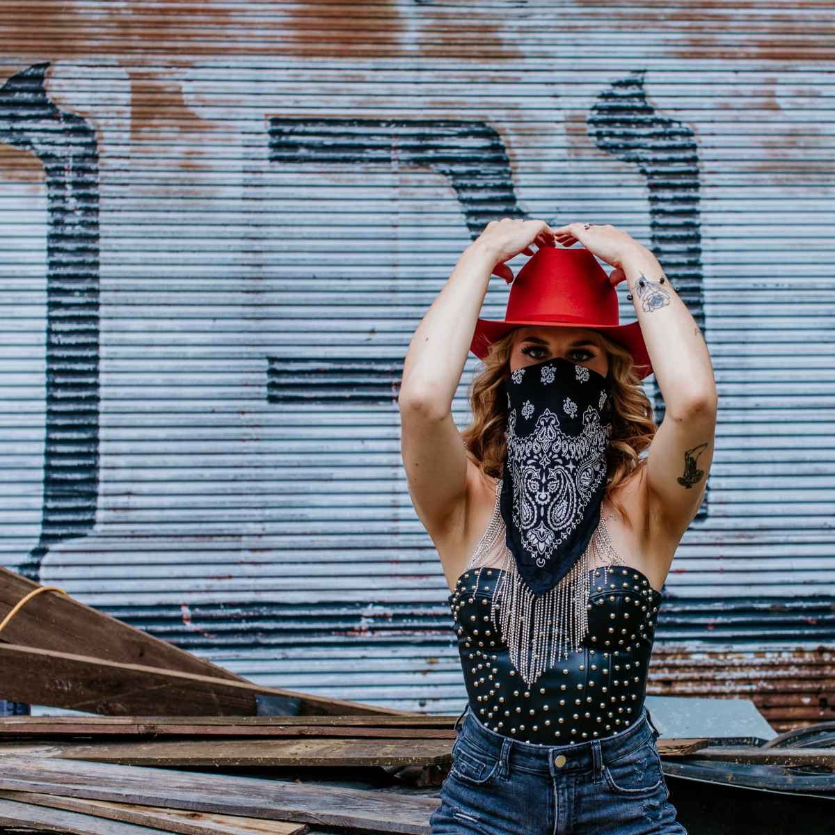 A person wearing a red hat and a black bandana stands in front of a corrugated metal background with graffiti.