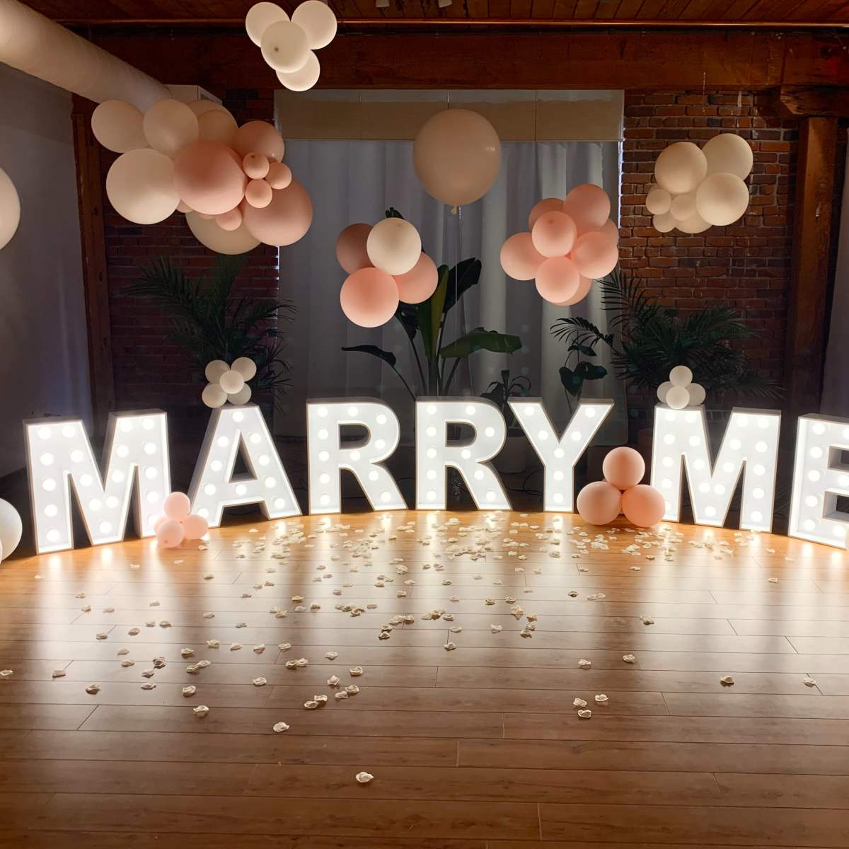 A decorative setup with a large illuminated "MARRY ME" sign surrounded by balloons and floral accents on a wooden floor.