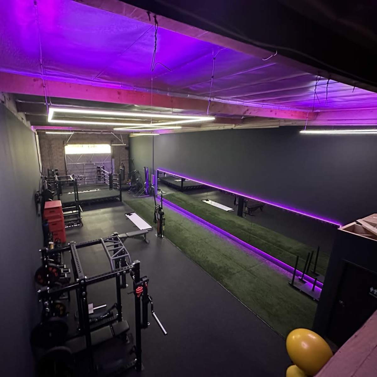 A modern gym with dark walls, purple lighting, and various workout equipment arranged along the floor.