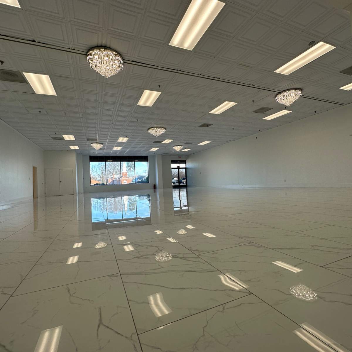 An empty commercial space with white walls, a high ceiling, and shiny marble flooring, illuminated by several ceiling lights and chandeliers.