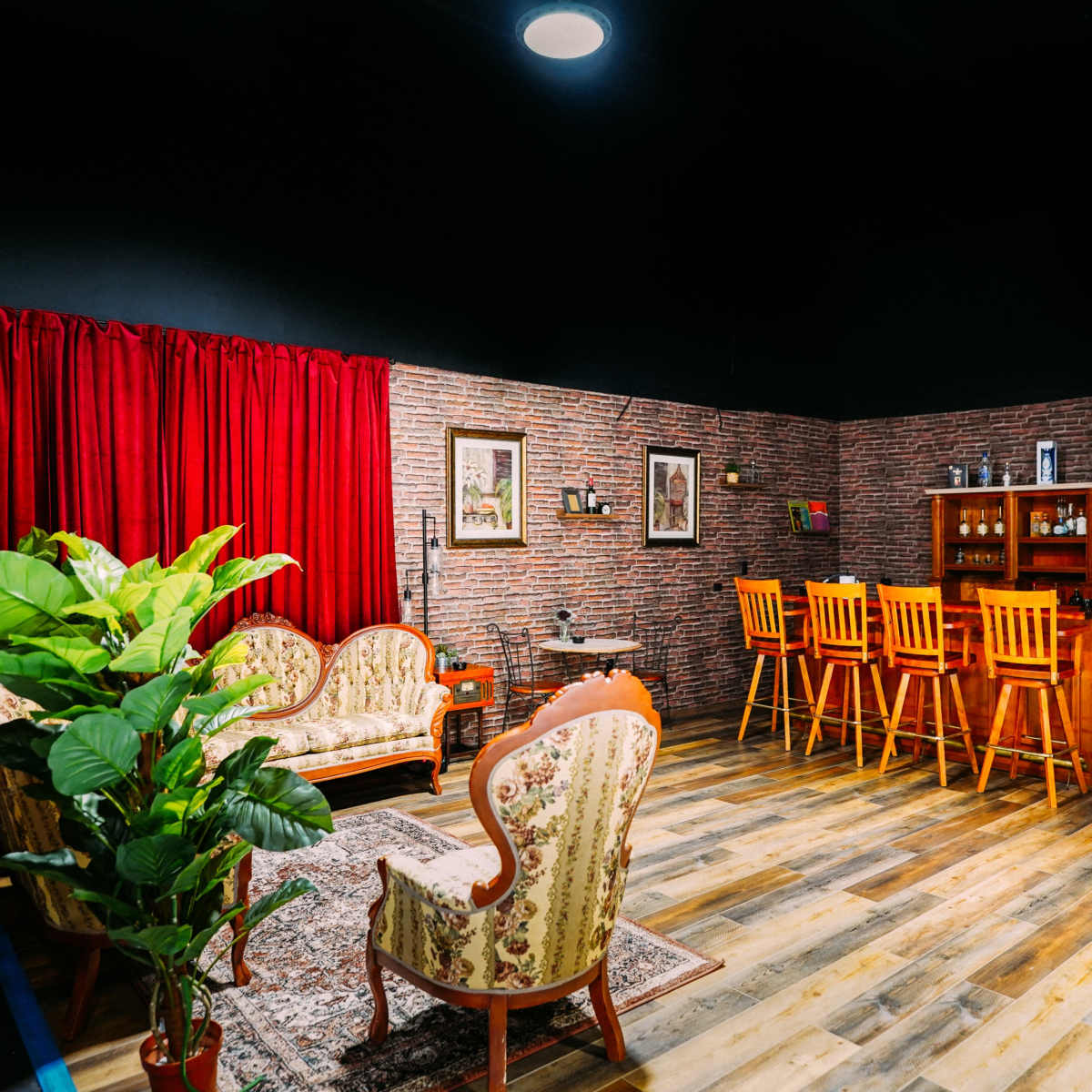 A cozy room with a brick wall, red curtains, a seating area with floral-patterned chairs, and a bar counter with wooden stools.